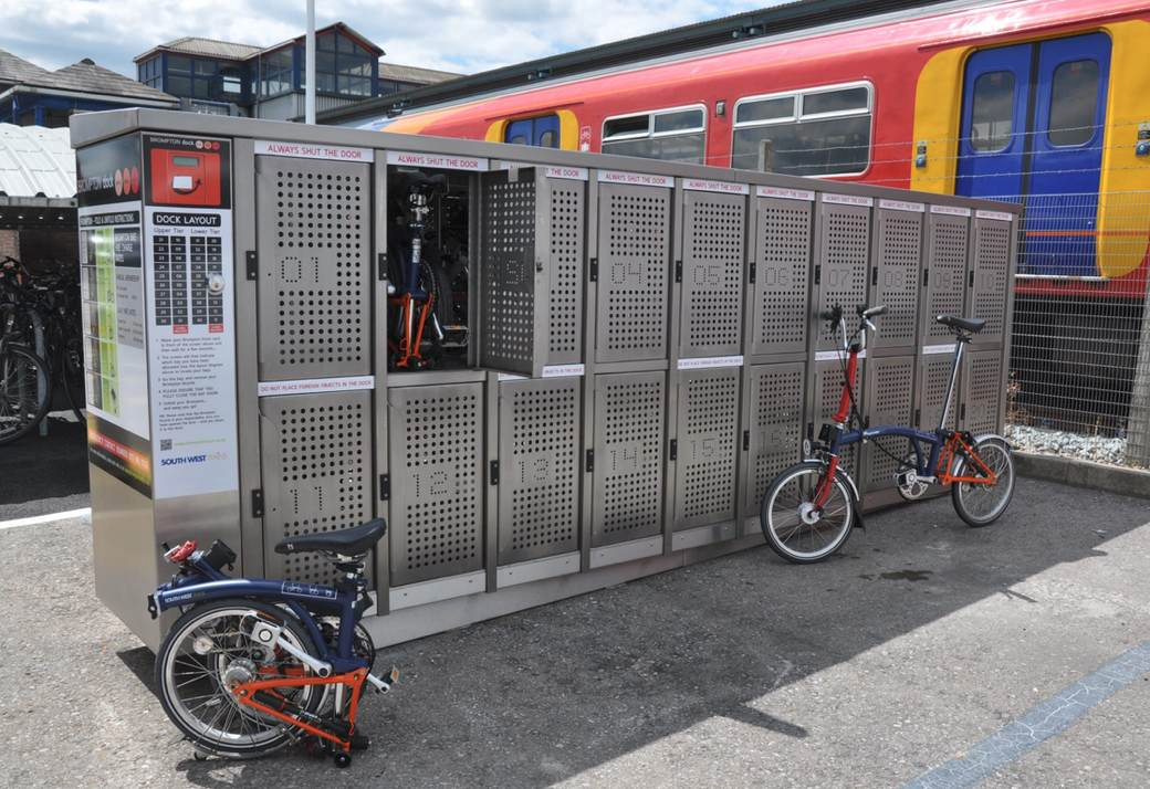 Casilleros de almacenamiento para bicicletas plegables en una estación de tren con bicicletas plegables estacionadas y un tren colorido en el fondo