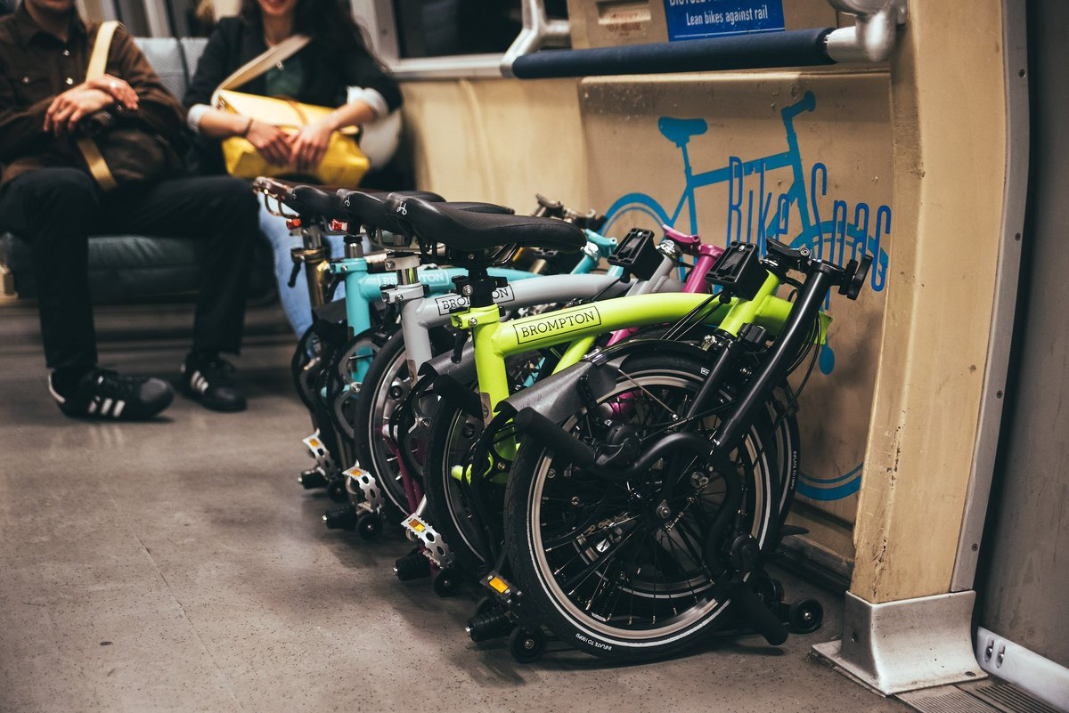 La imagen muestra el interior de un vagón de transporte público donde varias bicicletas plegables de la marca Brompton están organizadas en un espacio designado, etiquetado como "Bike Space". Las bicicletas están plegadas y colocadas ordenadamente, lo que permite ahorrar espacio y facilita su transporte en el tren. Al fondo, se observan dos pasajeros sentados, lo que sugiere una integración amigable de la movilidad en bicicleta con el transporte público. Esta disposición demuestra cómo el diseño de espacios en trenes puede acomodar de manera eficiente las necesidades de ciclistas urbanos.