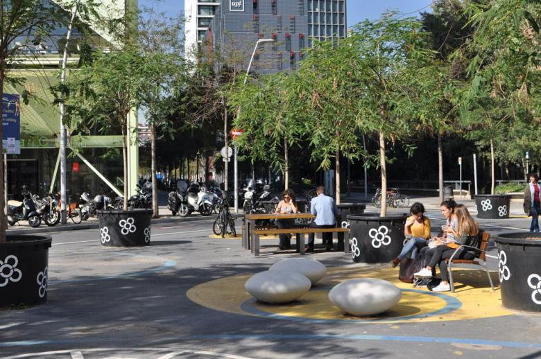 Espacio urbano (supermanzana) en Barcelona, al aire libre. Con árboles jóvenes en maceteros grandes negros decorados con flores blancas. Hay varias personas sentadas en bancos y en una mesa, socializando o usando sus dispositivos móviles. En el centro, un área circular amarilla y azul contiene varias estructuras de piedra redondeadas para sentarse. Al fondo, hay edificios modernos y un aparcamiento de bicicletas.