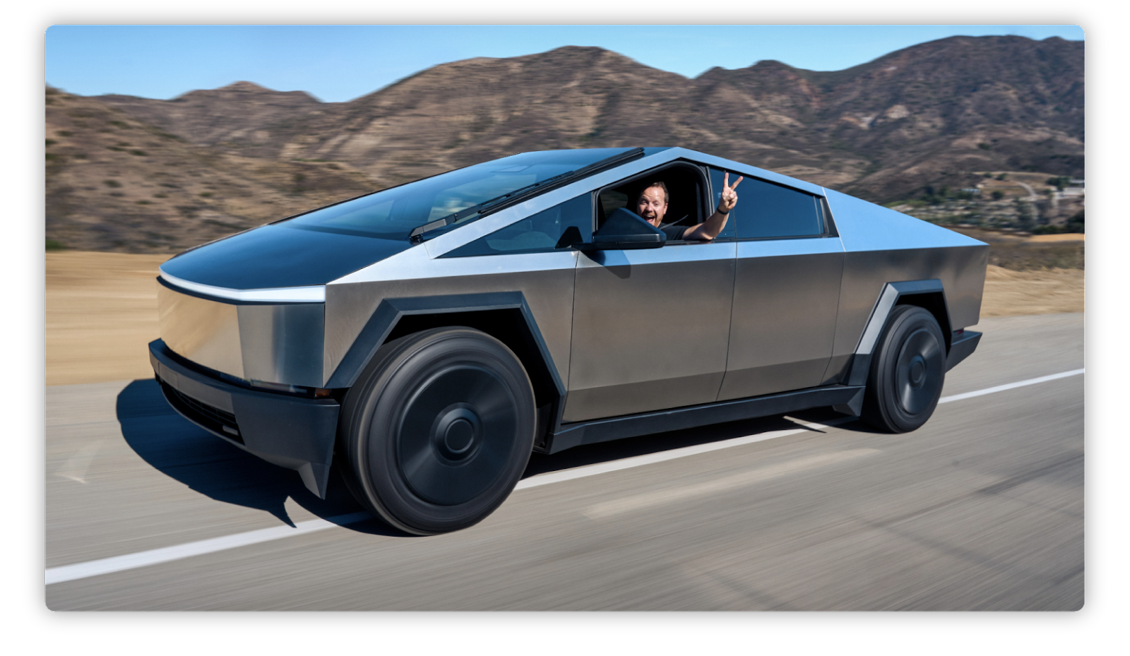 Imagen de una camioneta futurista de diseño angular y metálico, identificable como el Cybertruck de Tesla, circulando en una ruta. En el asiento del conductor, una persona sonriente saca la mano por la ventana y hace el signo de paz. Al fondo se observan montañas desérticas bajo un cielo despejado.
